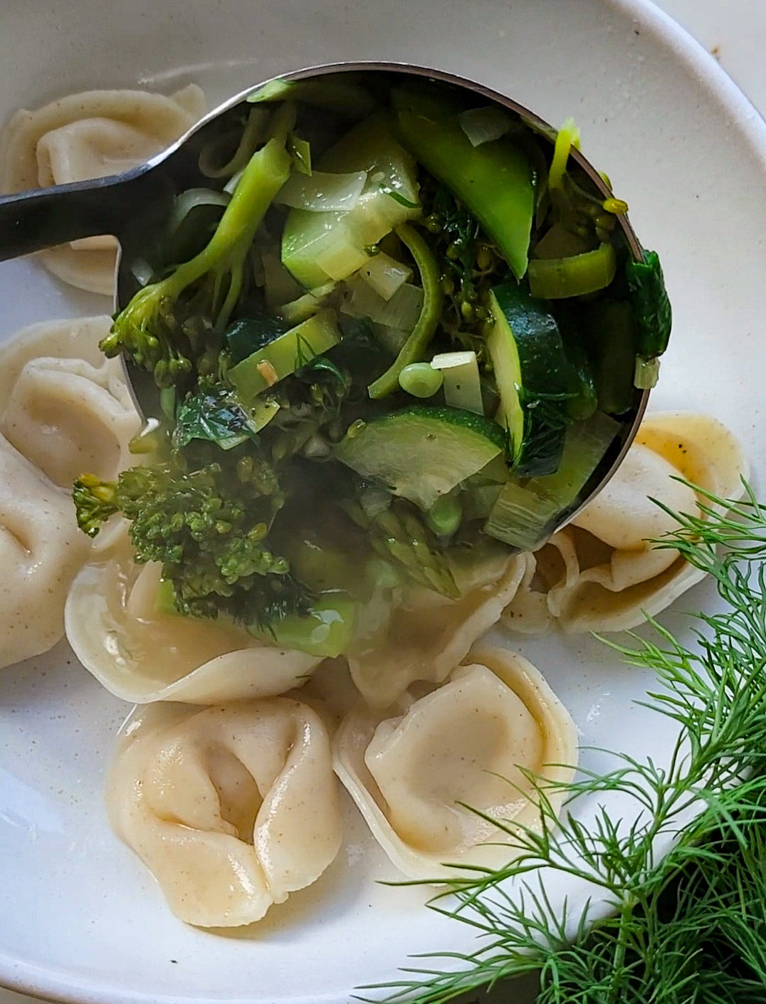 Pouring a broth with gently cooked broccoli, leeks, peas, asparagus and zucchini over cooked tortellini in a soup bowl.