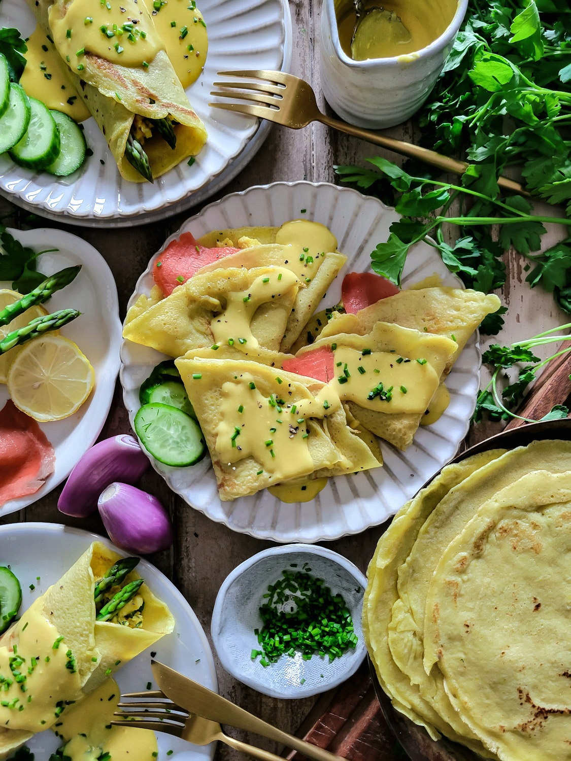 Plates of folded savory breakfast crepes, filled with asparagus, smoked salmon, creamy herbed scrambled eggs, hollandaise sauce, garnished with cucumbers and chives.