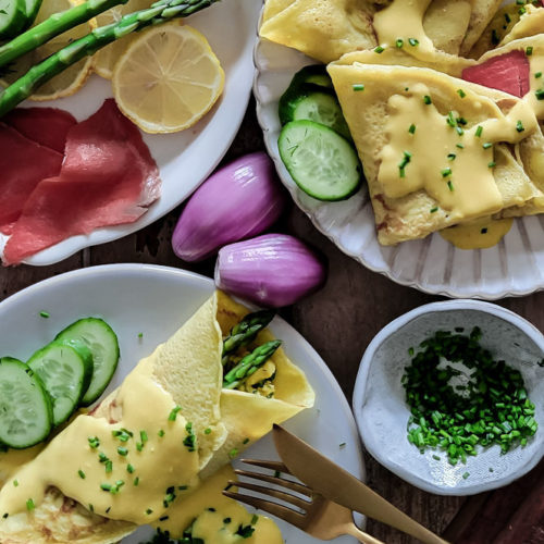 Plates of folded savory breakfast crepes, filled with asparagus, smoked salmon, creamy herbed scrambled eggs, hollandaise sauce, garnished with cucumbers and chives.