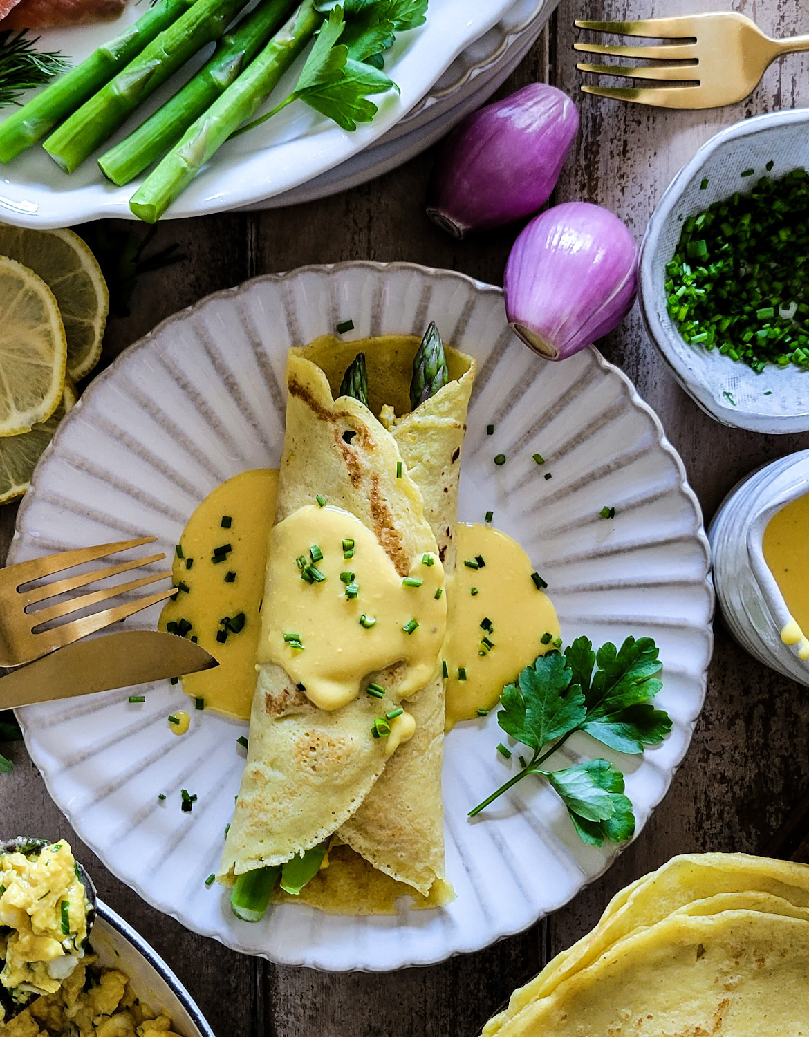 Plates of folded savory breakfast crepes, filled with asparagus, smoked salmon, creamy herbed scrambled eggs, hollandaise sauce, garnished with chives.