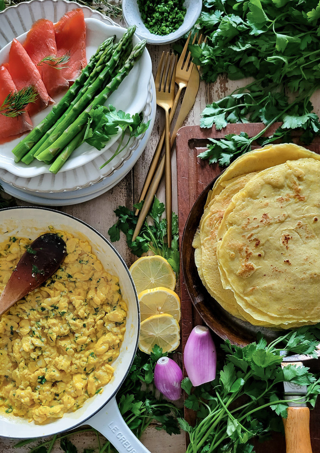Herb scrambled eggs in a skillet, stack of savory crepes in a crepe pan, smoked salmon and steamed asparagus
