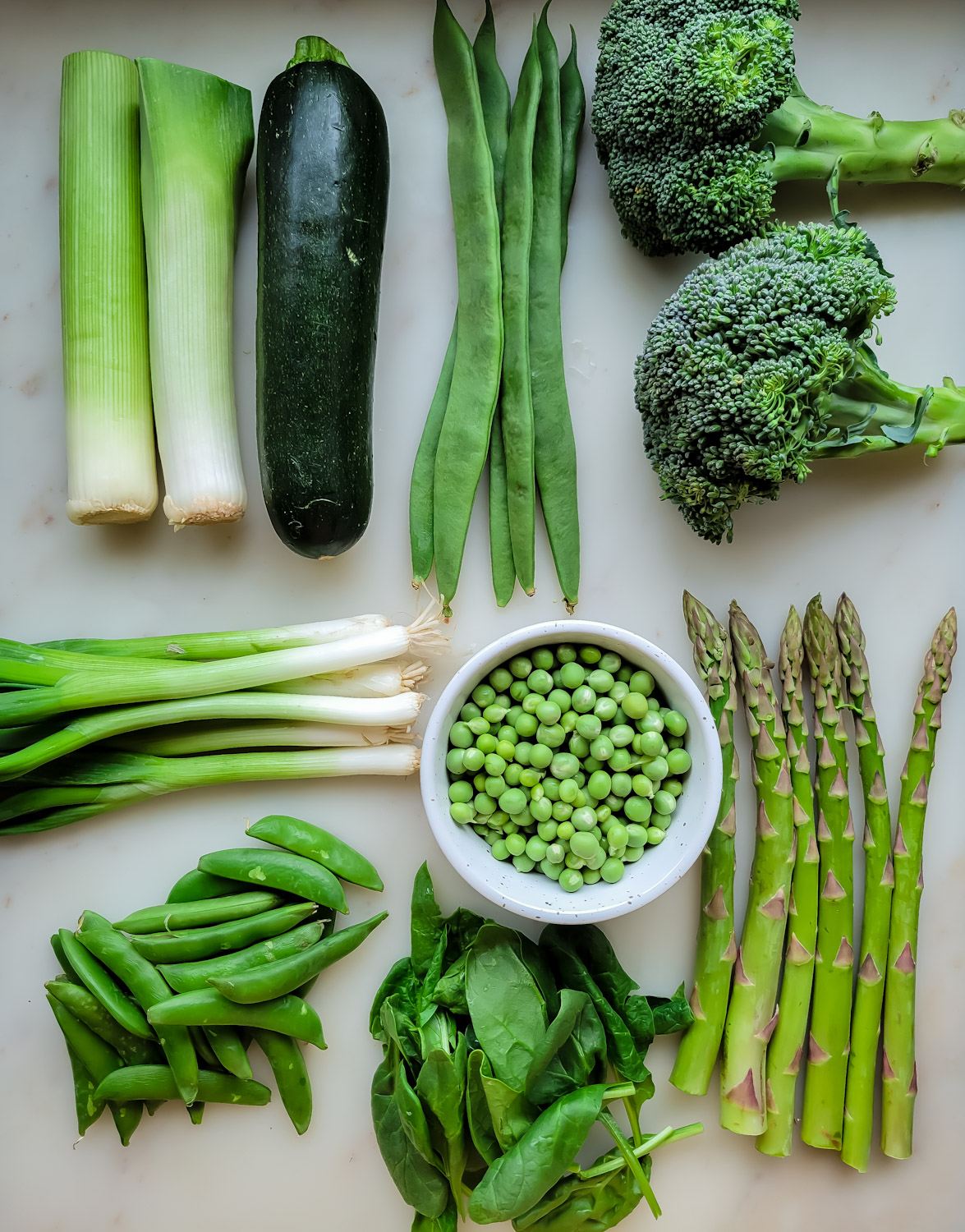 Fresh Spring Green vegetables are laid out in a grid. Leeks, zucchini, runner beans, broccoli, scallions, shelled peas, asparagus, snap peas and spinach