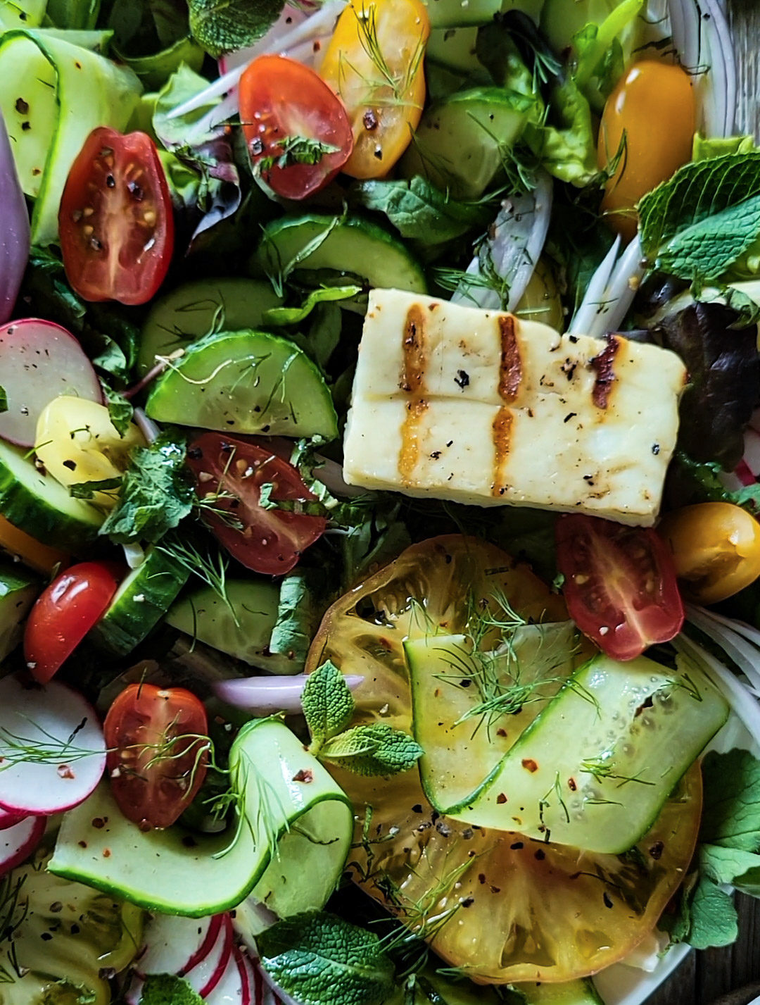 Close up of fattoush salad, cucumbers, tomatoes, onion, mint, radishes and topped with a piece of grilled halloumi cheese.