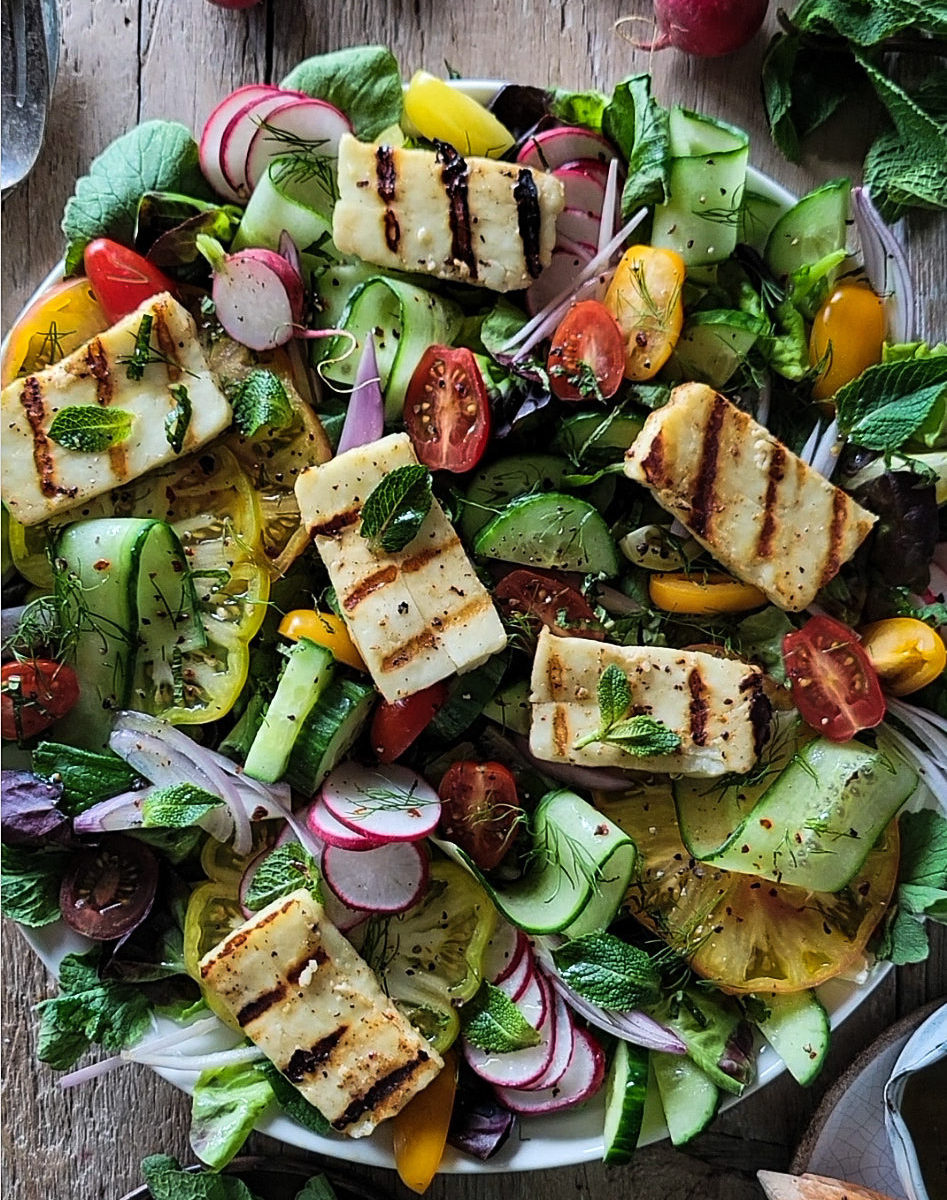 Platter of fattoush salad, cucumbers, tomatoes, onion, mint, radishes and slices of grilled halloumi cheese. There are radishes and mint leaves surrounding the platter, as well as serving utensils and small bowl of dressing.
