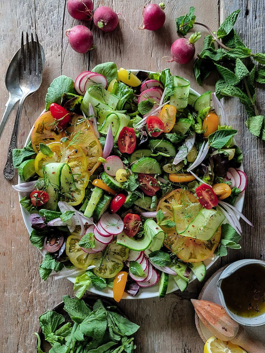 Platter of fattoush salad, cucumbers, tomatoes, onion, mint, and radishes. There are radishes and mint leaves surrounding the platter, as well as serving utensils and small bowl of dressing
