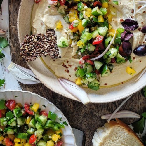 Bowl of creamy hummus covered with chopped cucumbers, peppers, onions, tomatoes and olives