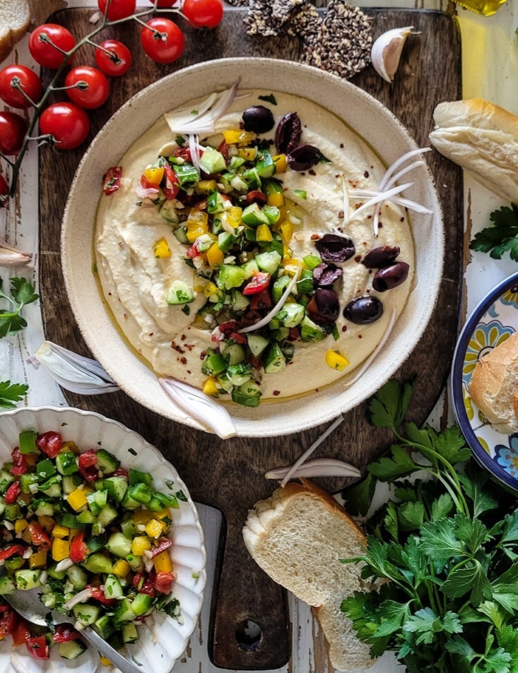 Bowl of creamy hummus covered with chopped cucumbers, peppers, onions, tomatoes and olives, surrounded by tomatoes on the vine, crackers and parsley