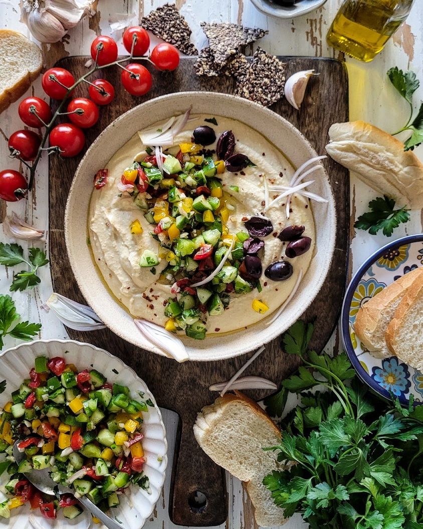 Bowl of creamy hummus covered with chopped cucumbers, peppers, onions, tomatoes and olives, surrounded by tomatoes on the vine, crackers and parsley