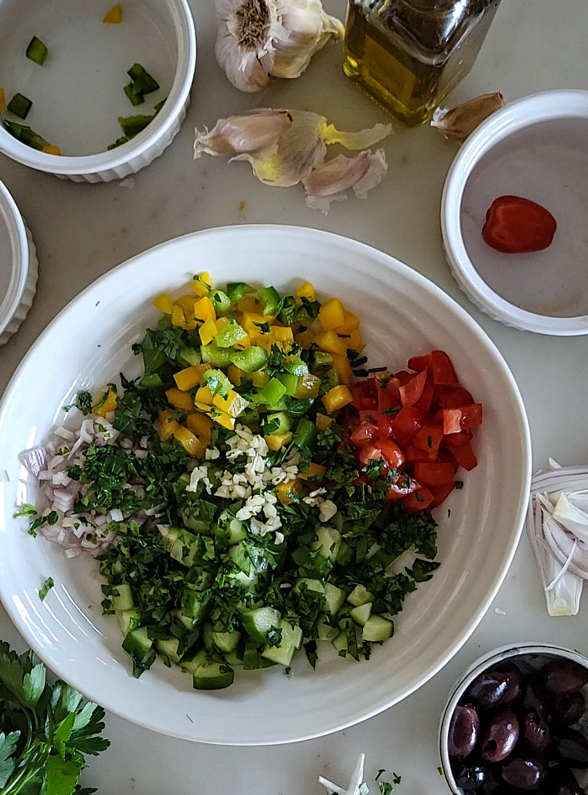 Bowl of chopped cucumbers, peppers, onions, and tomatoes, a bowl of olives, garlic cloves and olive oil to the side,