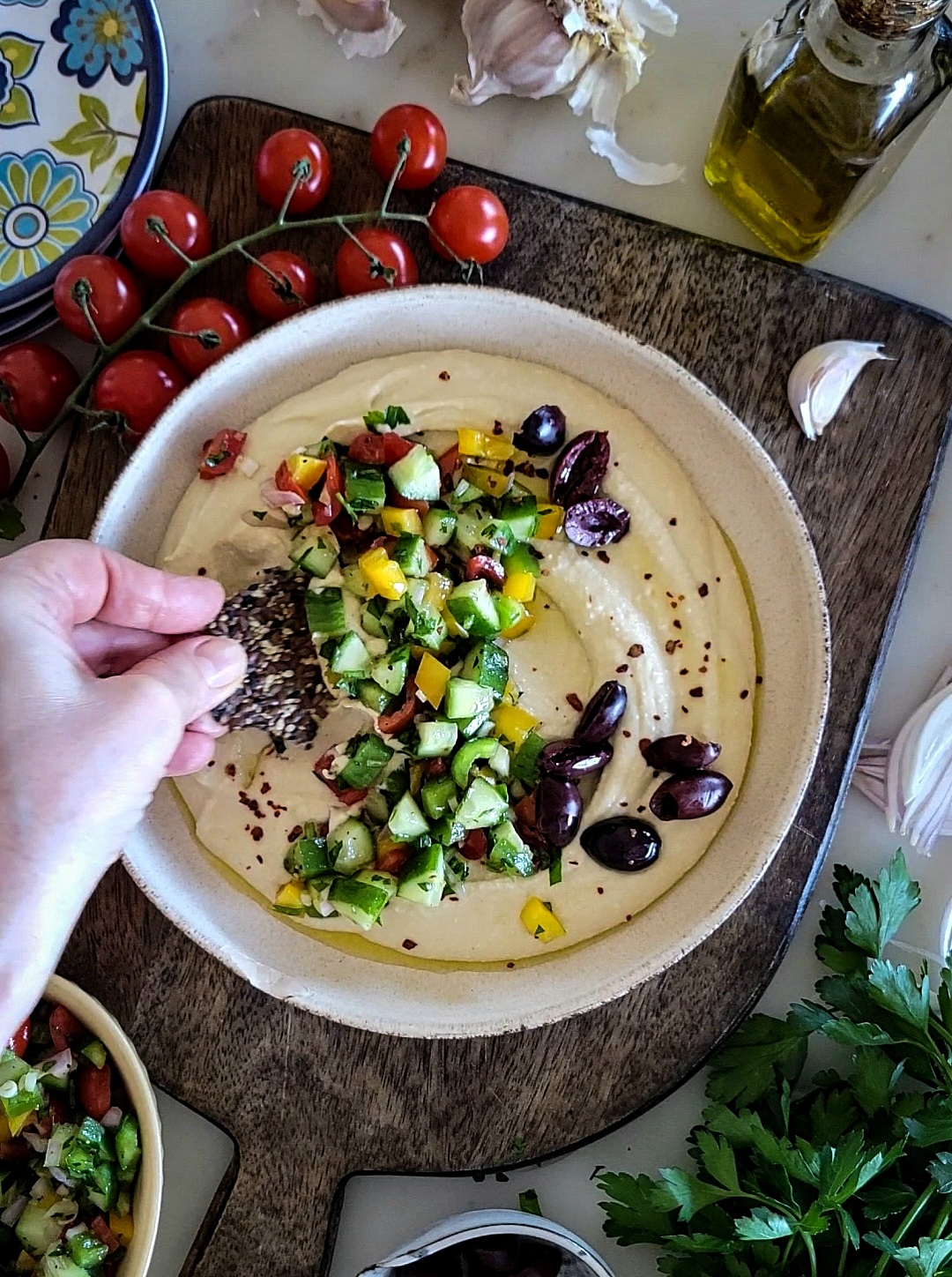 Bowl of creamy hummus covered with chopped cucumbers, peppers, onions, tomatoes and olives, surrounded by tomatoes on the vine, crackers and parsley