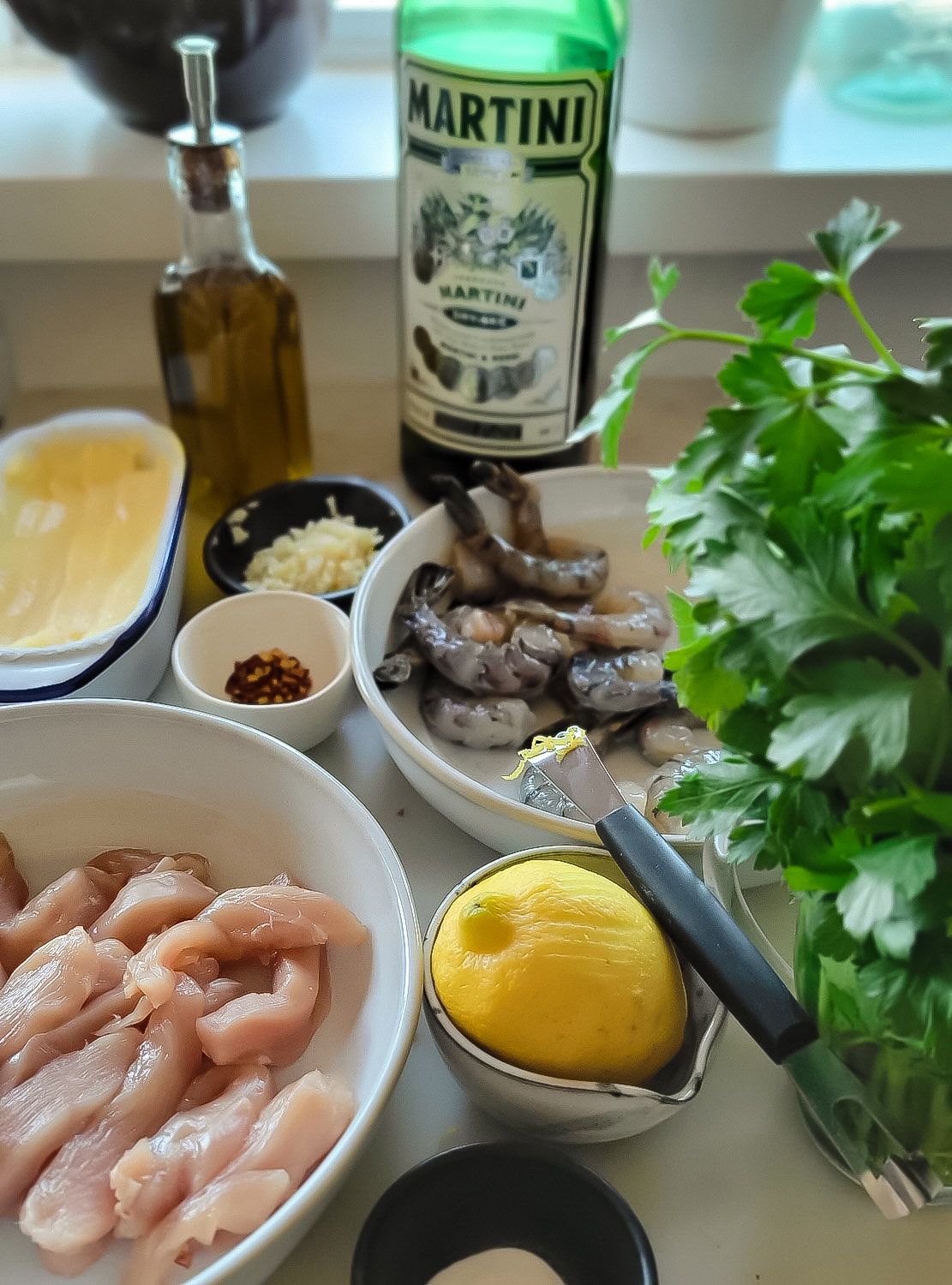 a bowl of peeled shrimp and a bowl of chicken breast cut into strips are surrounded by parsley, lemon, chilli flakes, garlic, butter and a bottle of dry vermouth