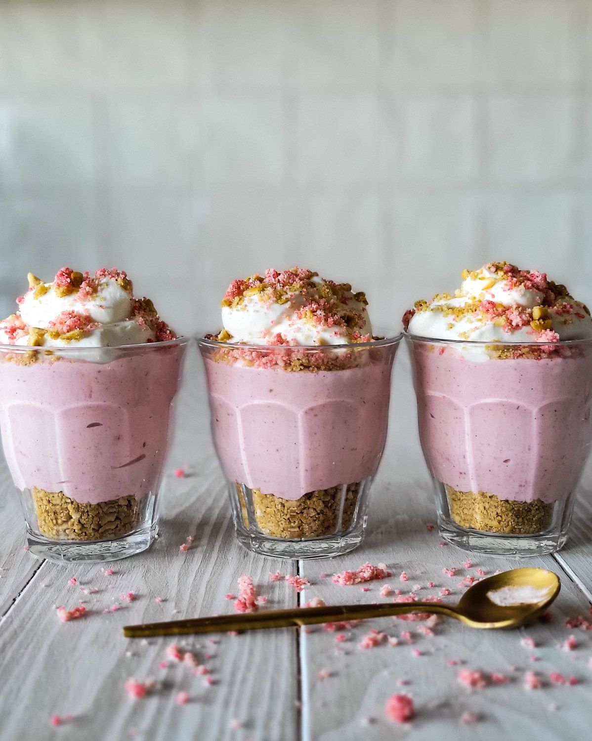 Three small glasses containing a pretzel base, strawberry mousse, and topped with whipped cream, strawberry shortcake crumble and more pretzel crumble. There are crumbs and a spoon on the table