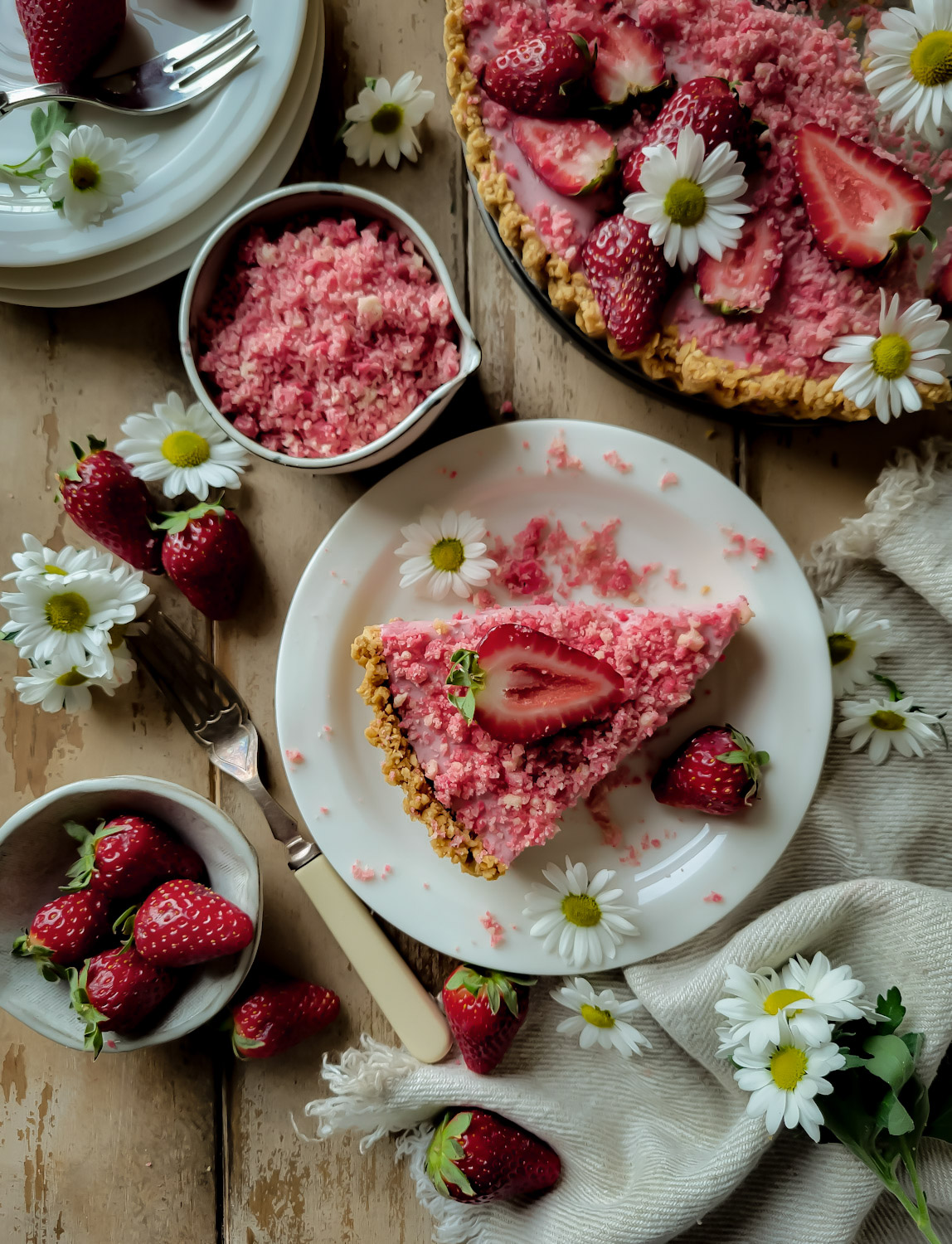 A slice of strawberry shortcake mousse tart on a plate with shortcake crumble in a small bowl next to it, daises and fresh strawberries to the side.