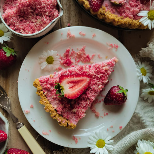 A slice of strawberry shortcake mousse tart on a plate with shortcake crumble in a small bowl next to it, daises and fresh strawberries to the side.