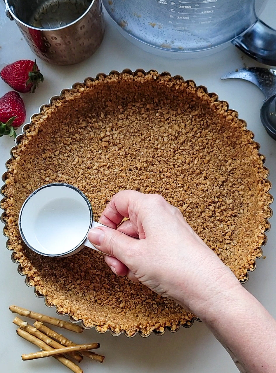 hand holding a measuring cup smoothing the pretzel crust into the tart pan