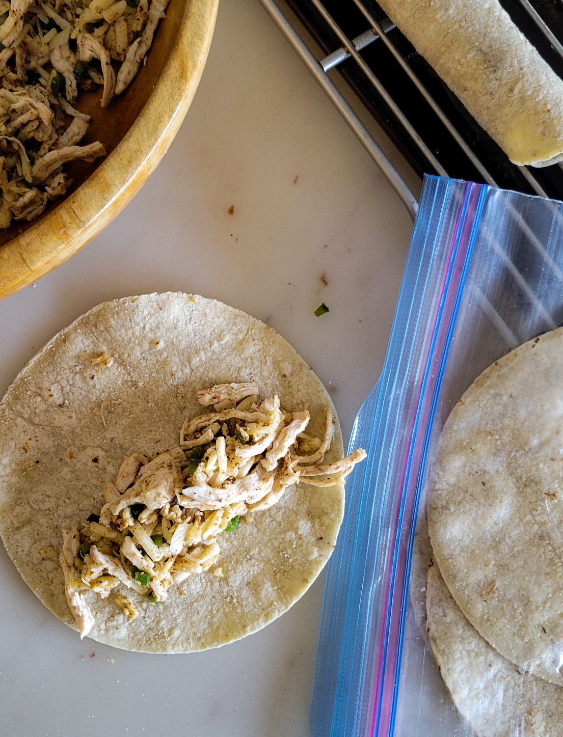 filling a corn tortilla with chicken and cheese to roll into a taquito, freezer bag of warm tortillas to the side