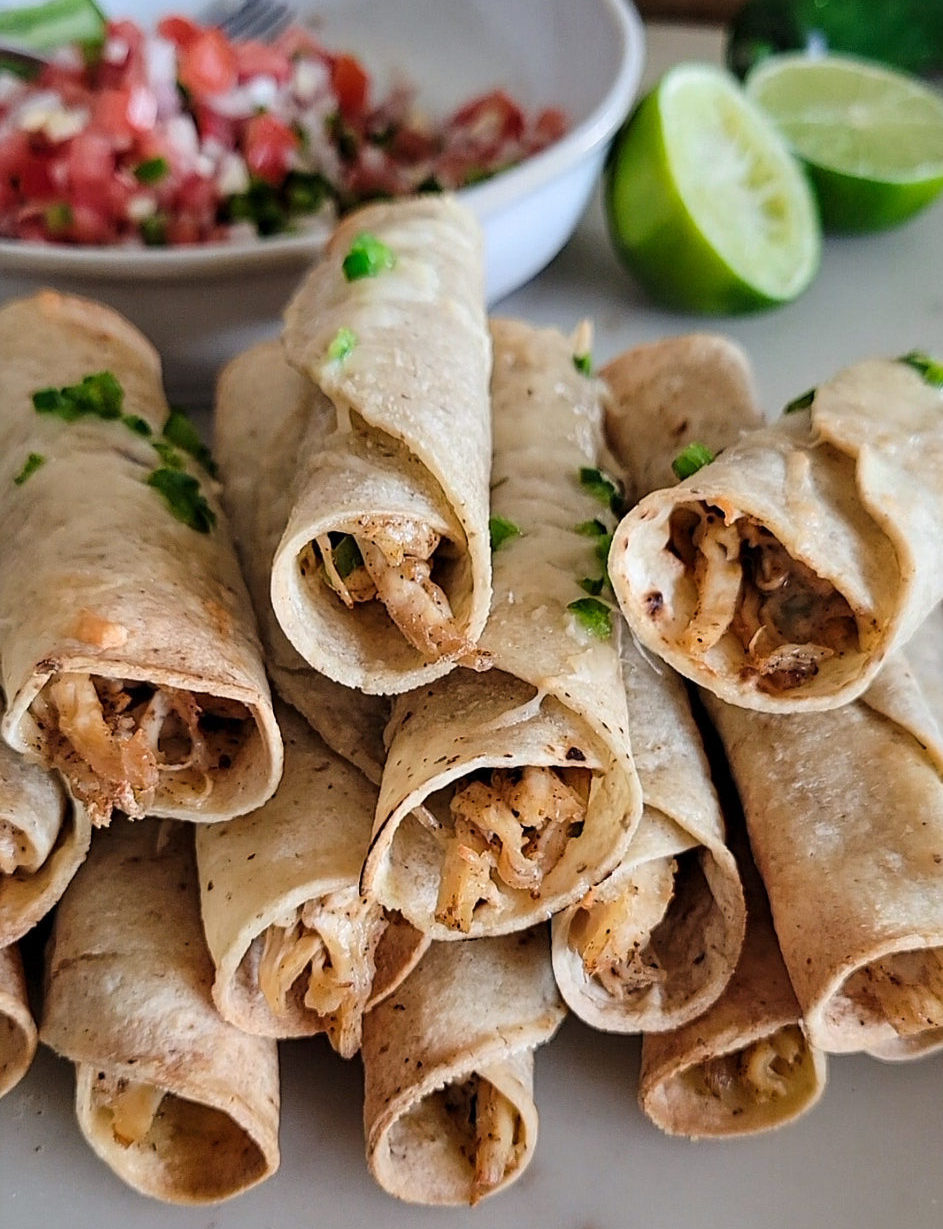 A pile of baked chicken taquitos with a bowl of pico de gallo in the background