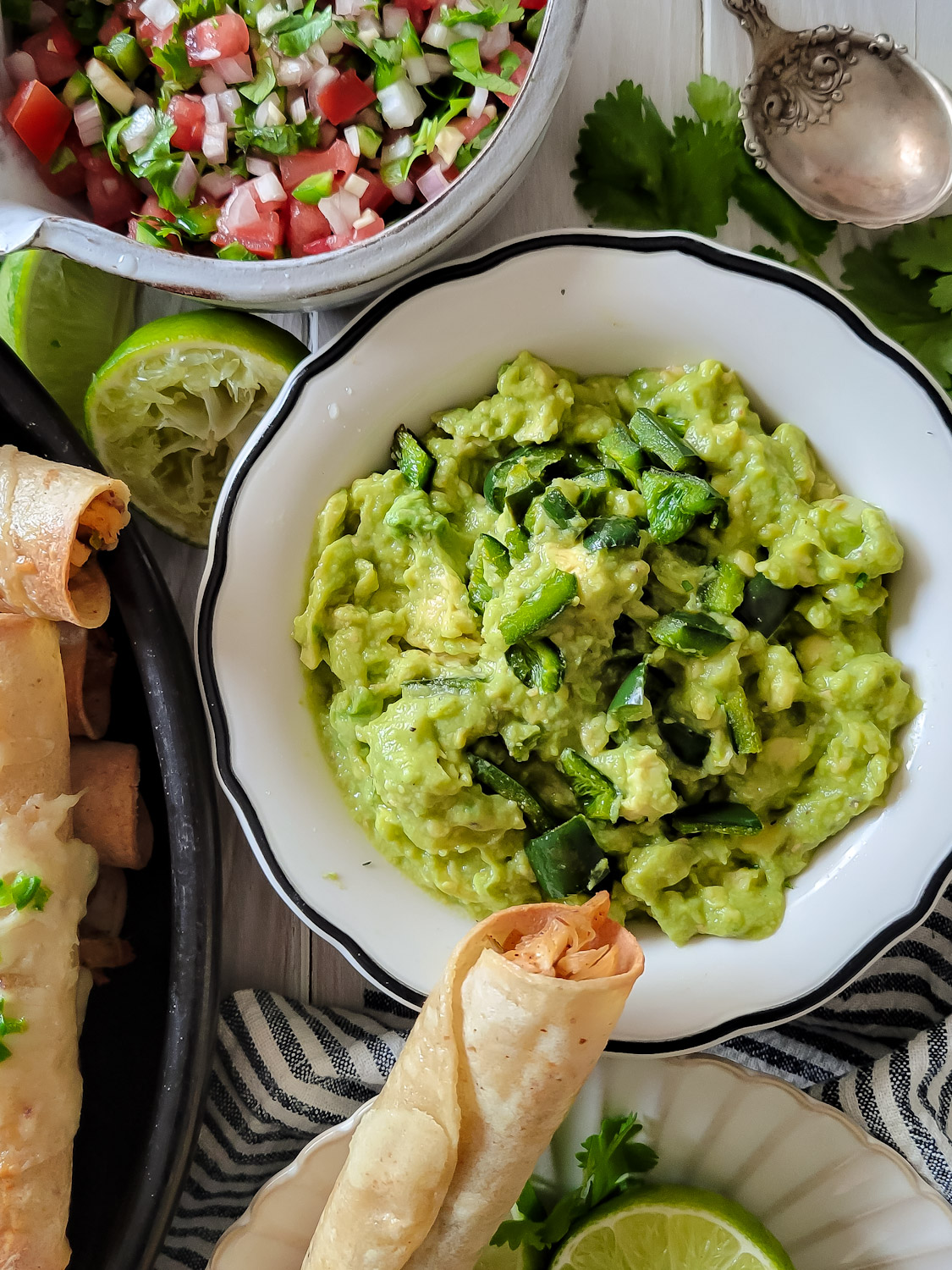 a bowl of classic guacamole with the addition of chopped roasted poblano pepper. Pico de gallo can be seen in the background, and a taquito is sitting on the edge of the bowl.