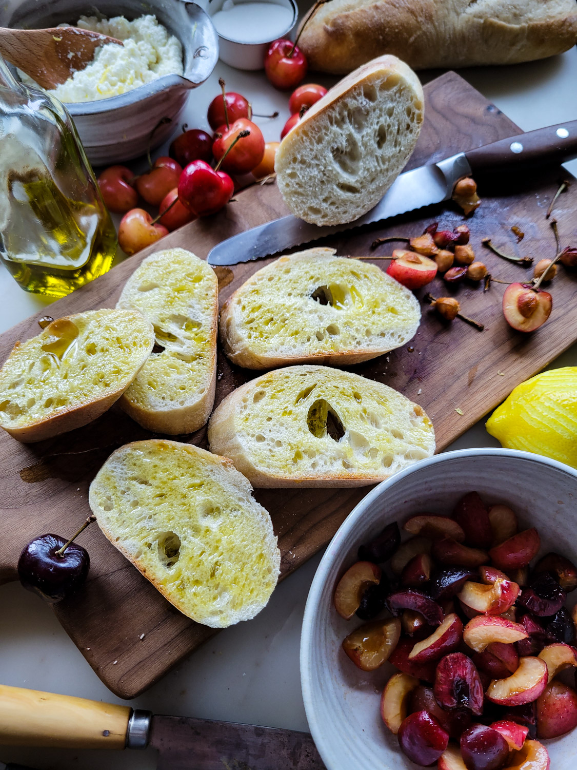 prep shot of sliced mini baguette, chopped cherries, a bottle of olive oil and a bowl of lemon ricotta cheese