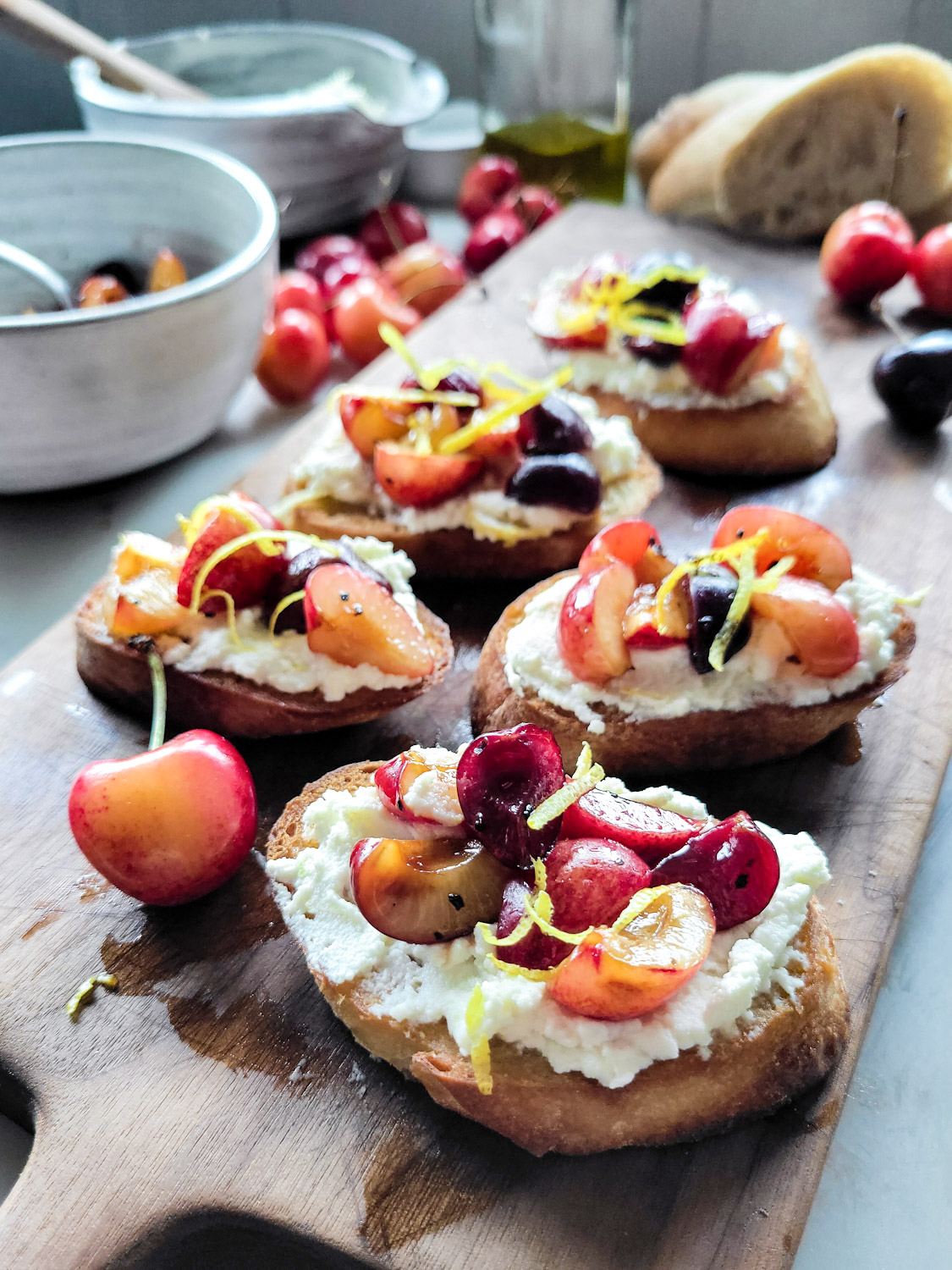 A wooden cutting board with toasted crostini slices spread with lemon ricotta cheese and topped with macerated chopped cherries
