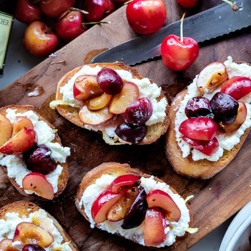 Toasted crostini slices spread with lemon ricotta cheese and topped with macerated chopped cherries sitting on a cutting board