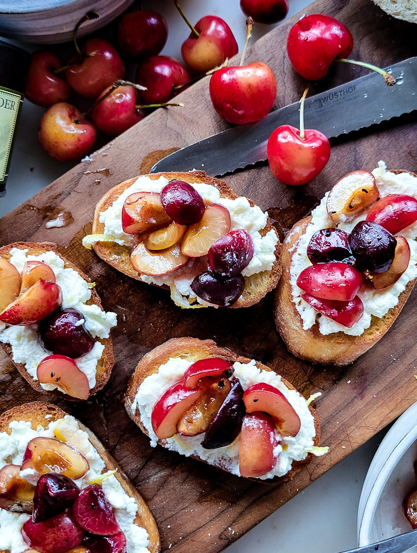 Toasted crostini slices spread with lemon ricotta cheese and topped with macerated chopped cherries sitting on a cutting board