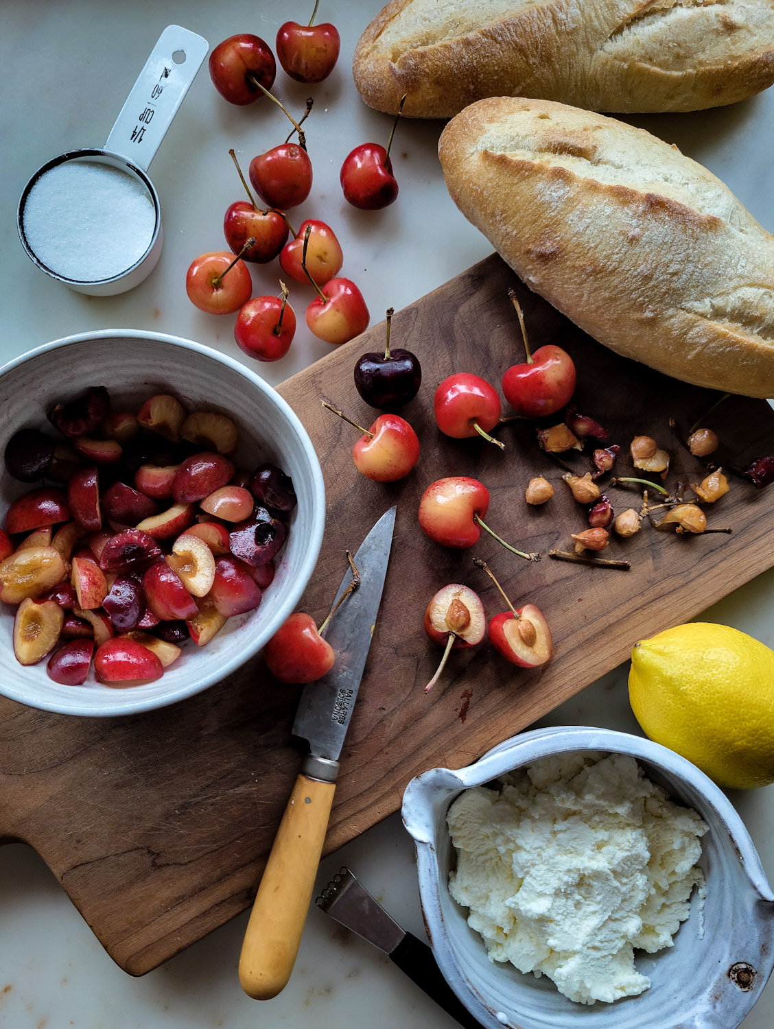Prep shot of a cutting board, macerated chopped cherries, crusty rolls, lemon and a bowl of ricotta cheese