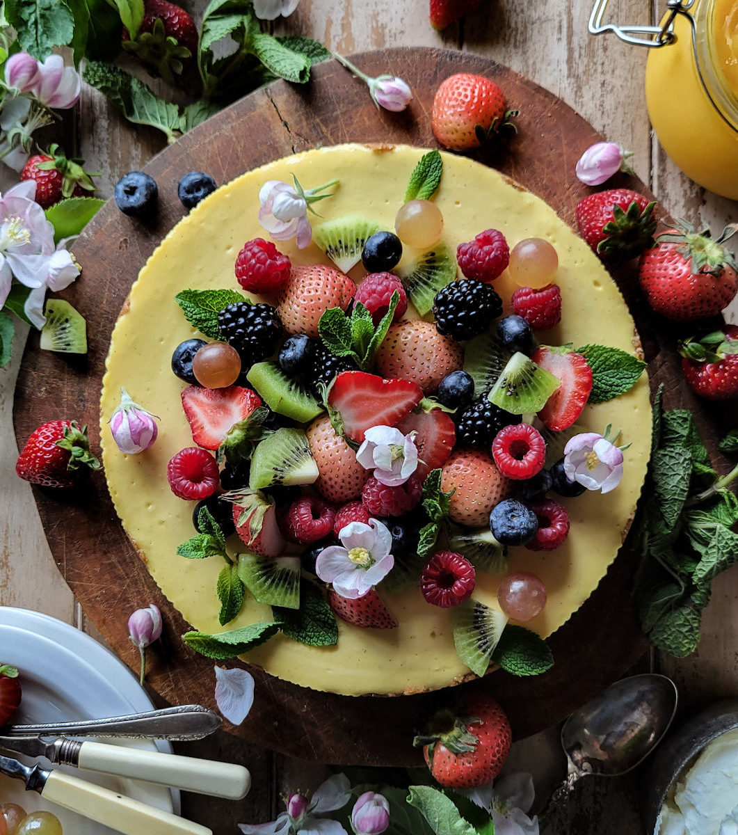 lemon cheesecake topped with fresh berries, mint leaves and apple blossoms, surrounded by more apple blossoms and lemon curd