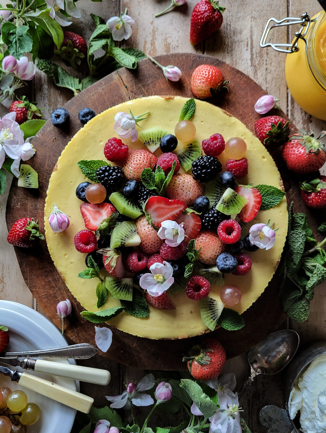 lemon cheesecake topped with fresh berries, mint leaves and apple blossoms, surrounded by more apple blossoms and lemon curd