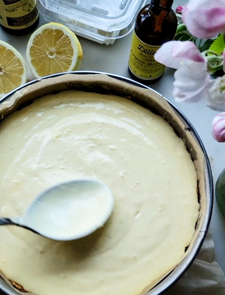 Lemon Cheesecake batter in a springform pan with lemons and a bottle of lemon bitters in the background