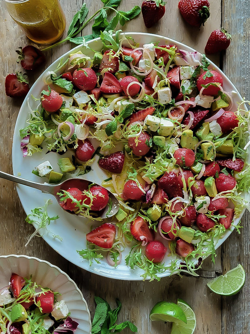 Serving plate of watermelon, strawberry and avocado salad with feta and lime vinaigrette. A smaller plate, mint leaves, strawberries, and lime wedges are next to it.