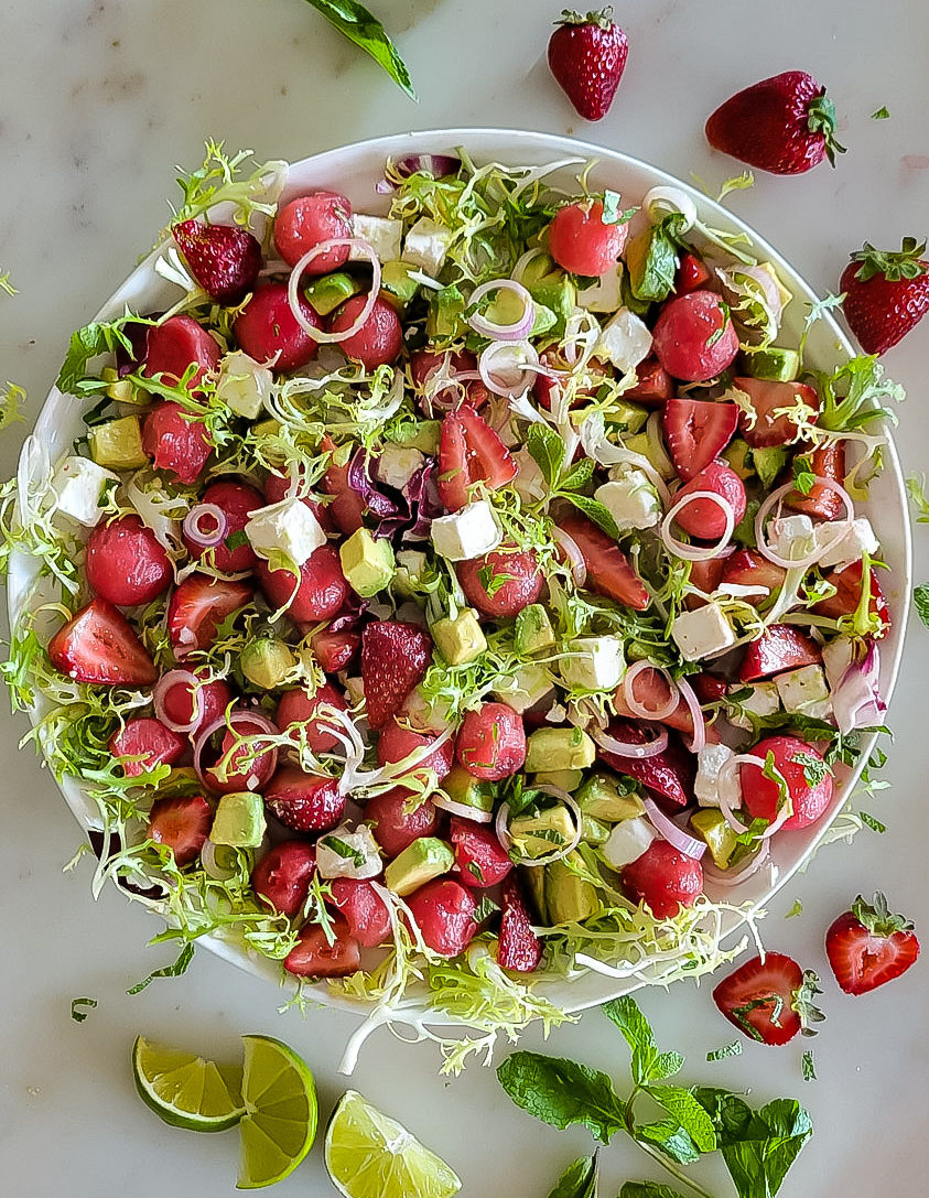 A platter with watermelon balls, strawberries, feta cheese, avocado and frisee lettuce, with lime wedges, mint and strawberries scattered around it.