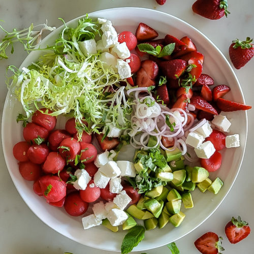 A platter with watermelon balls, strawberries, feta cheese, avocado and frisee lettuce, with lime wedges, mint and strawberries scattered around it.