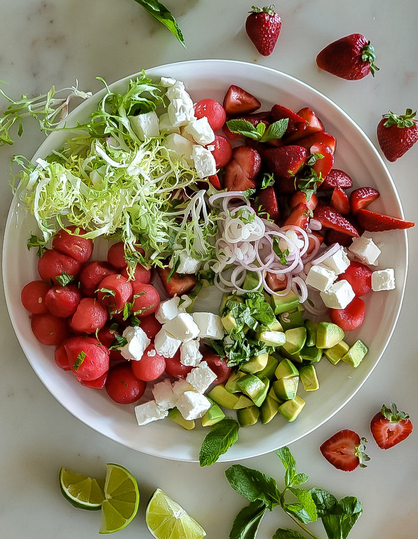 A platter with watermelon balls, strawberries, feta cheese, avocado and frisee lettuce, with lime wedges, mint and strawberries scattered around it.