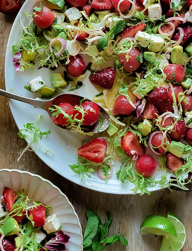 Serving plate of watermelon, strawberry and avocado salad with feta and lime vinaigrette. A smaller plate, mint leaves and lime wedges are next to it.