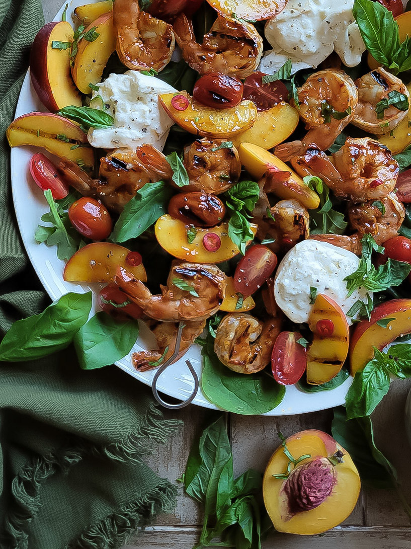 A caprese salad on a platter with grilled glazed shrimp, peaches and tomatoes, with fresh basil.