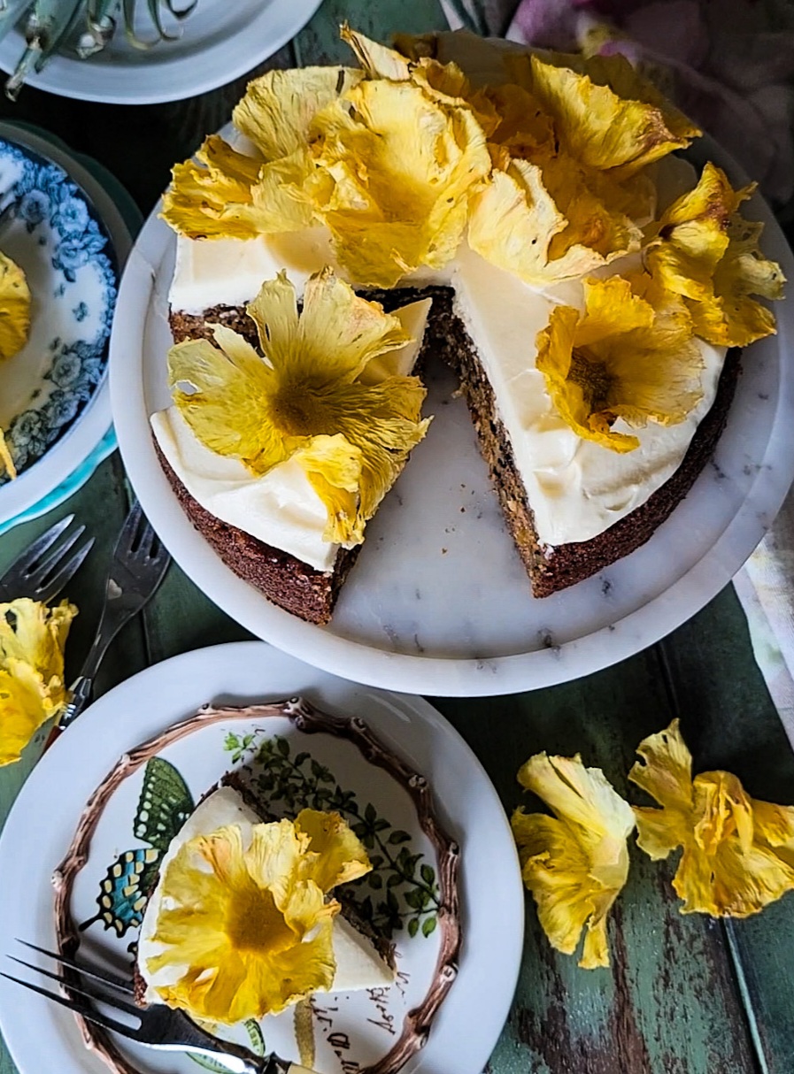 Zucchini Carrot Cake with cream cheese icing and pienapple flowers, one slice sitting on a plate next to the cake.