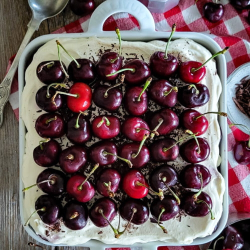 Baking dish filled with chocolate layered Black Forest Tiramisu, surrounded by fresh cherries and a red checkered cloth
