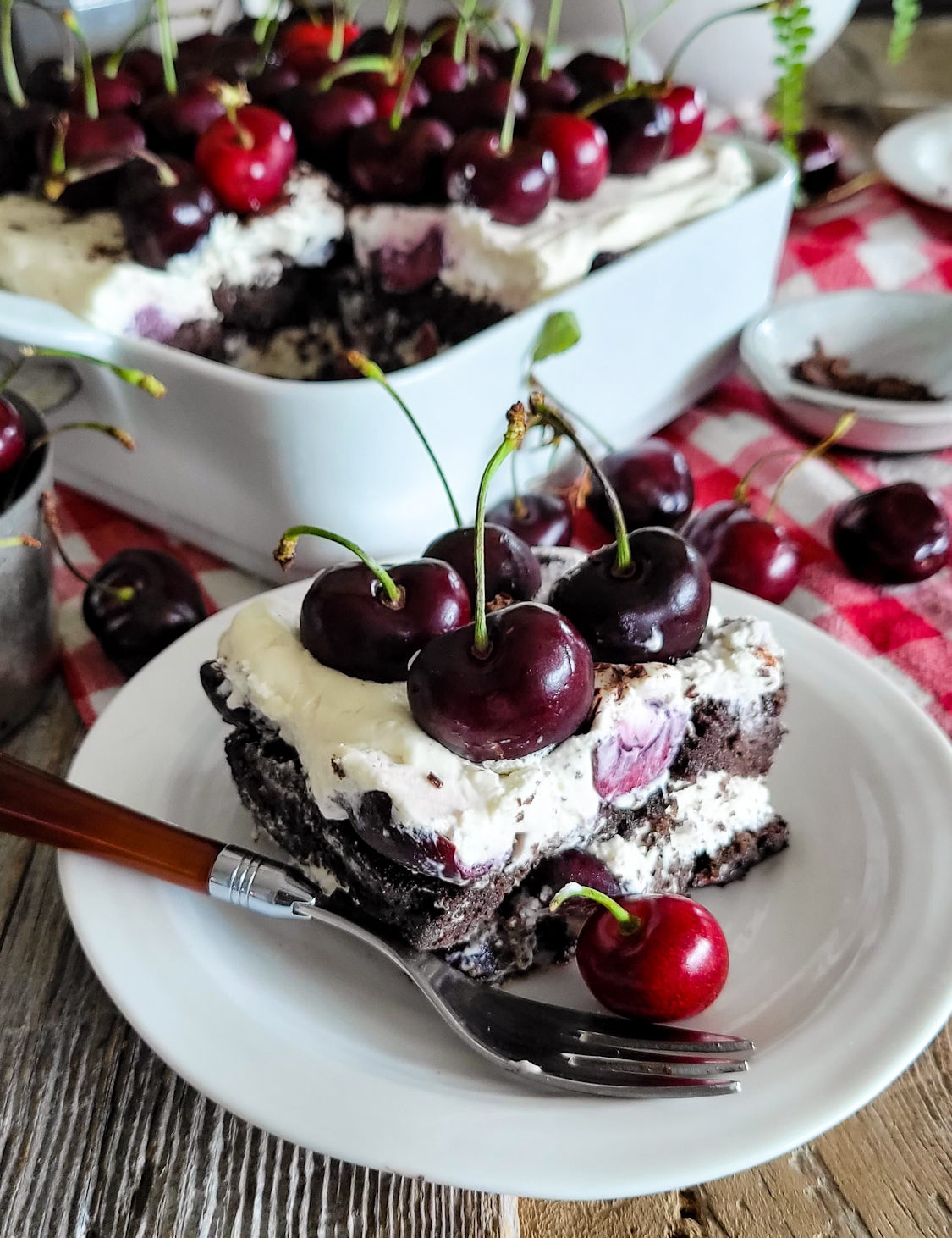 Closeup of a slice of Black Forest Tiramisu, topped with fresh cherries. The dish and pan of dessert are surrounded by fresh cherries, and everything is sitting on a red checkered cloth.