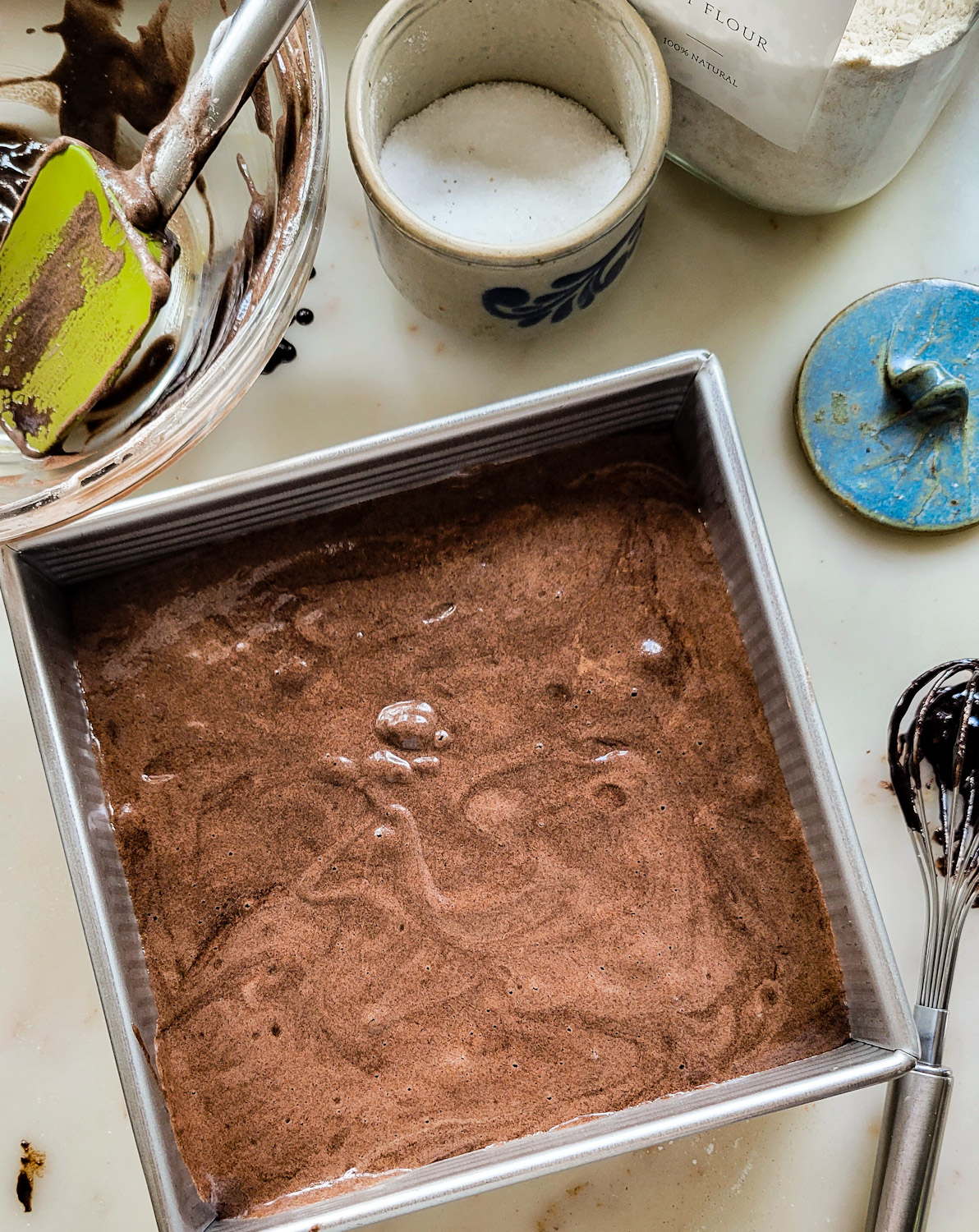 pan filled with chocolate sponge cake batter for black forest tiramisu. Pan is surrounded by mixing bowl, battered covered whisk, and baking ingredients.