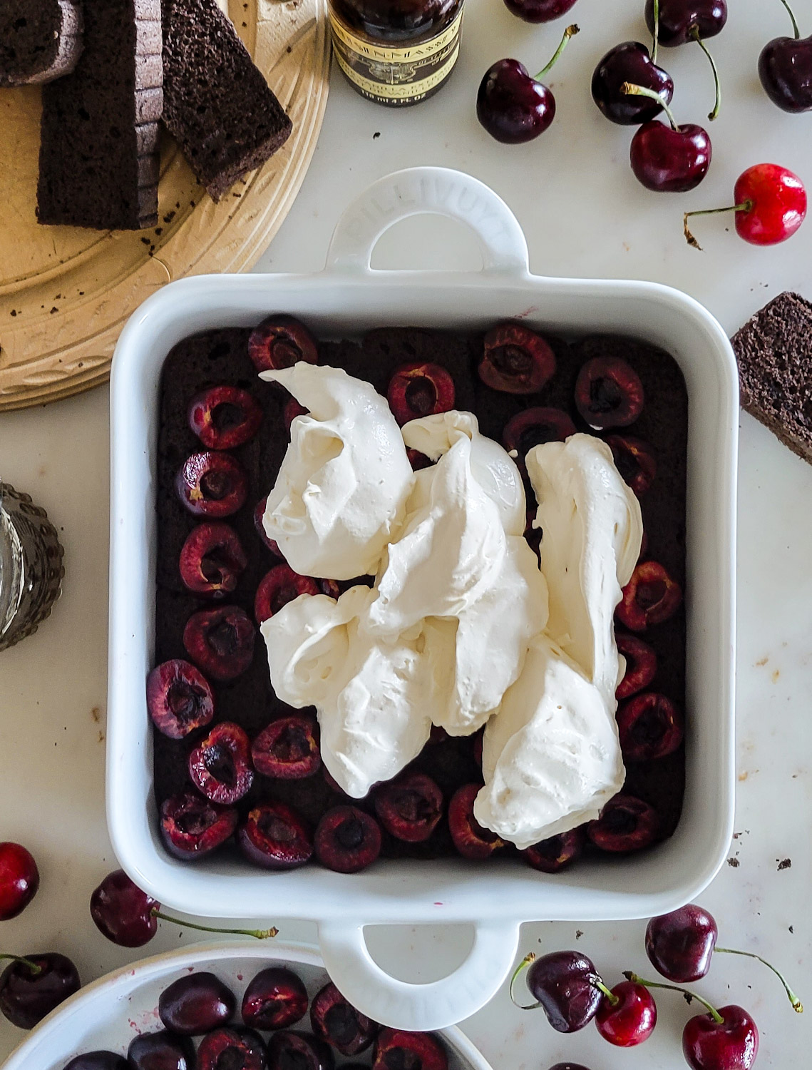 baking dish with chocolate sponge cake layer, fresh cherry layer, and mascarpone whipped cream layer, for a Black Forest Tiramisu. Surrounded by fresh cherries and sliced sponge cake.