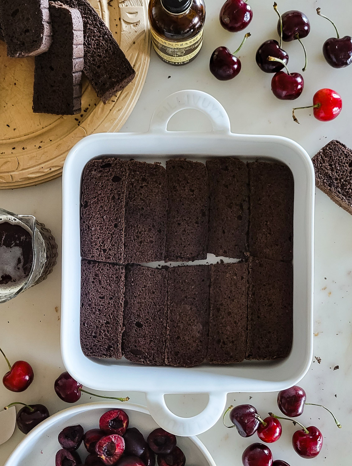 Baking dish with chocolate sponge cake layered for Black forest Tiramisu. Pan is surrounded by fresh cherries and more sponge cake slices.