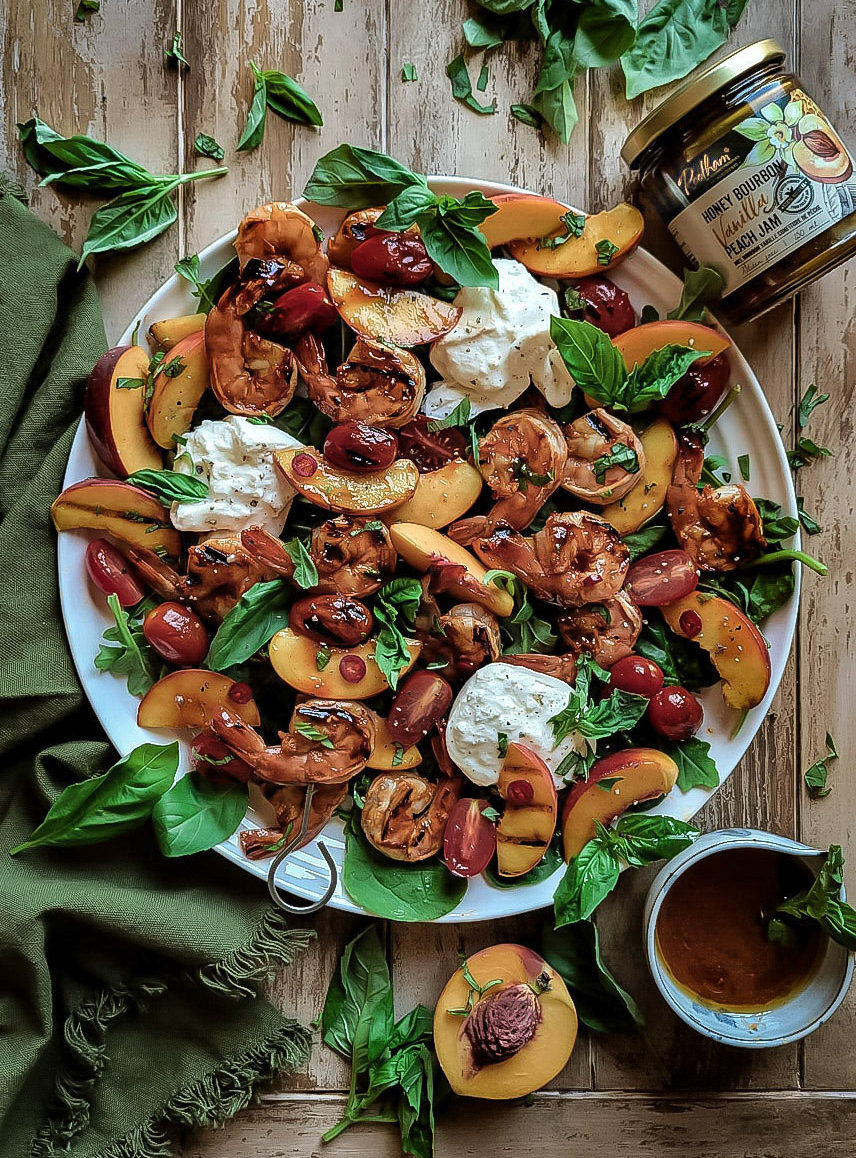 Platter of peach and sambal oelek glazed shrimp, peaches and tomatoes set into greens, burrata cheese and basil leaves. The jar of peach jam, basil leaves and a sliced peach surround the platter.