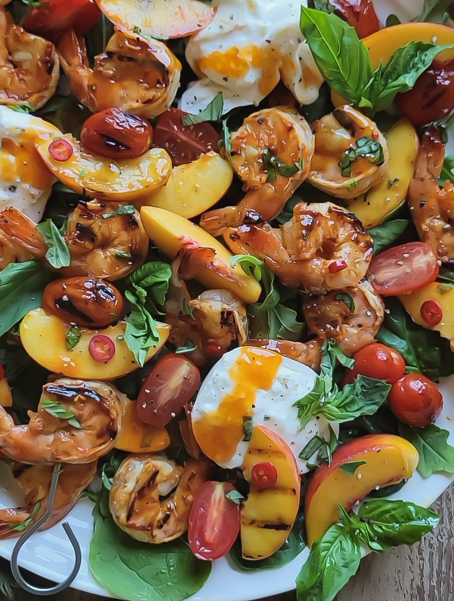 close up of grilled shrimp, peaches and tomatoes sitting on greens with burrata and basil