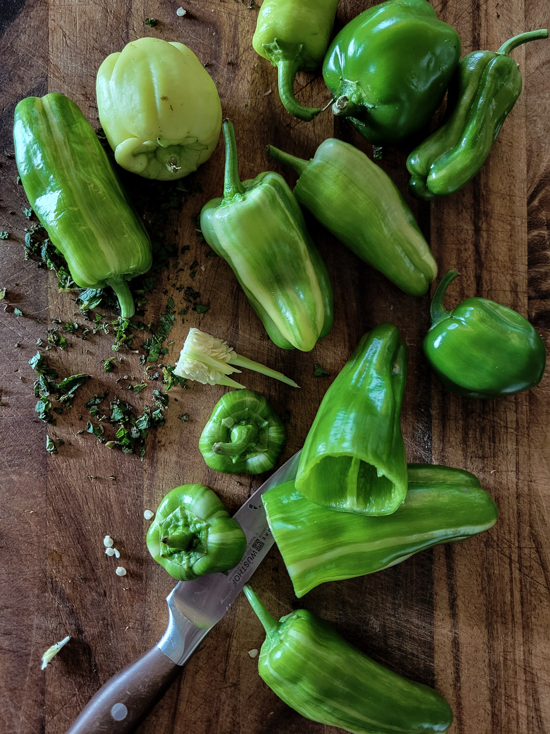 Cutting board with mini green peppers being prepped to be stuffed. A knife and chopped mint are also on the cutting board.
