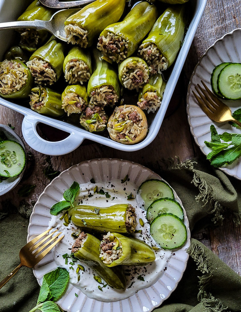 A baking dish filled with lamb and rice stuffed mini peppers. A plate with cumin yogurt, several stuffed peppers, pine nuts and mint is ready to be eaten.