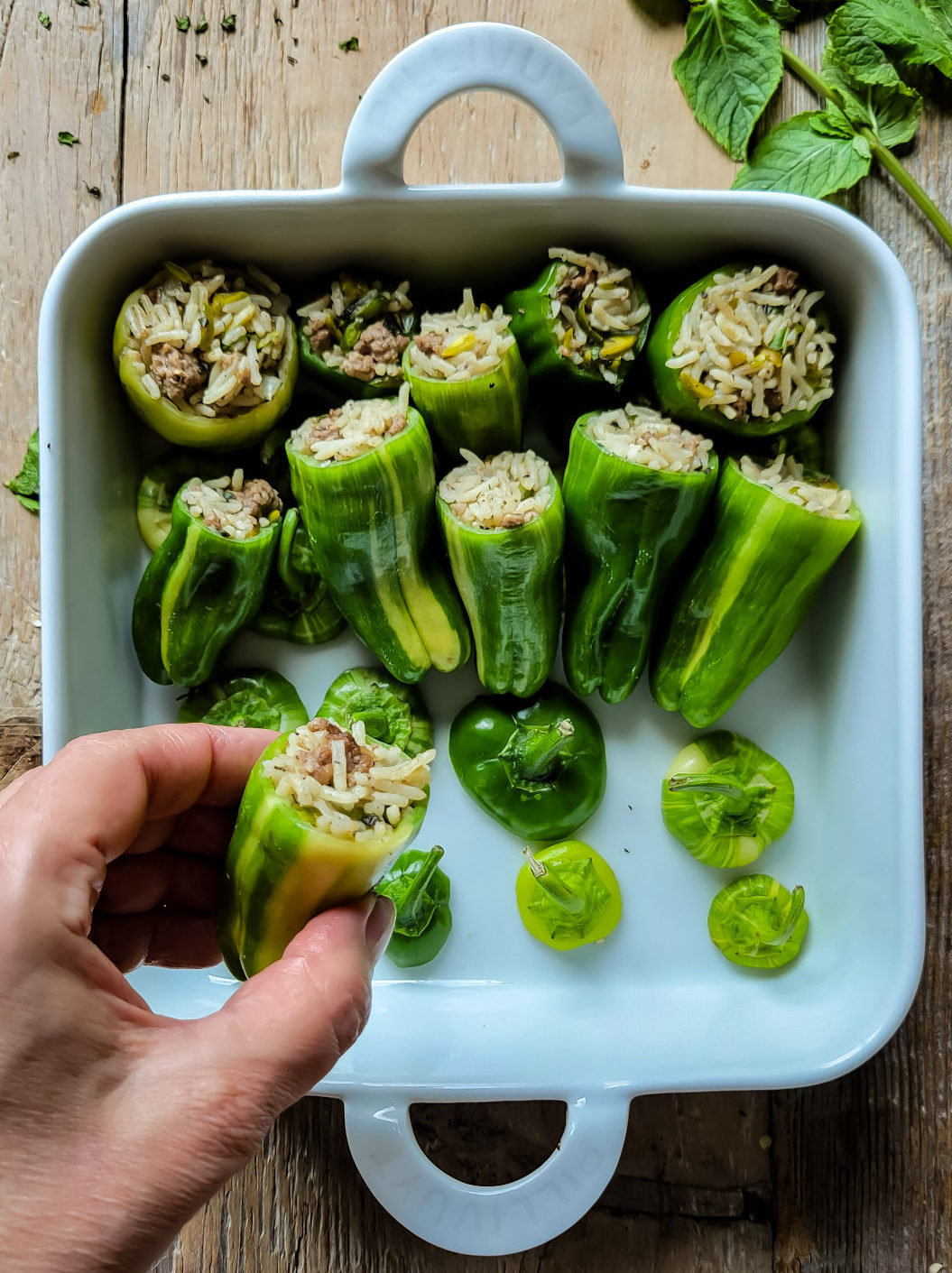 A baking dish is being filled with lamb and rice stuffed peppers getting ready to go in the oven. Mint leaves are on the side.