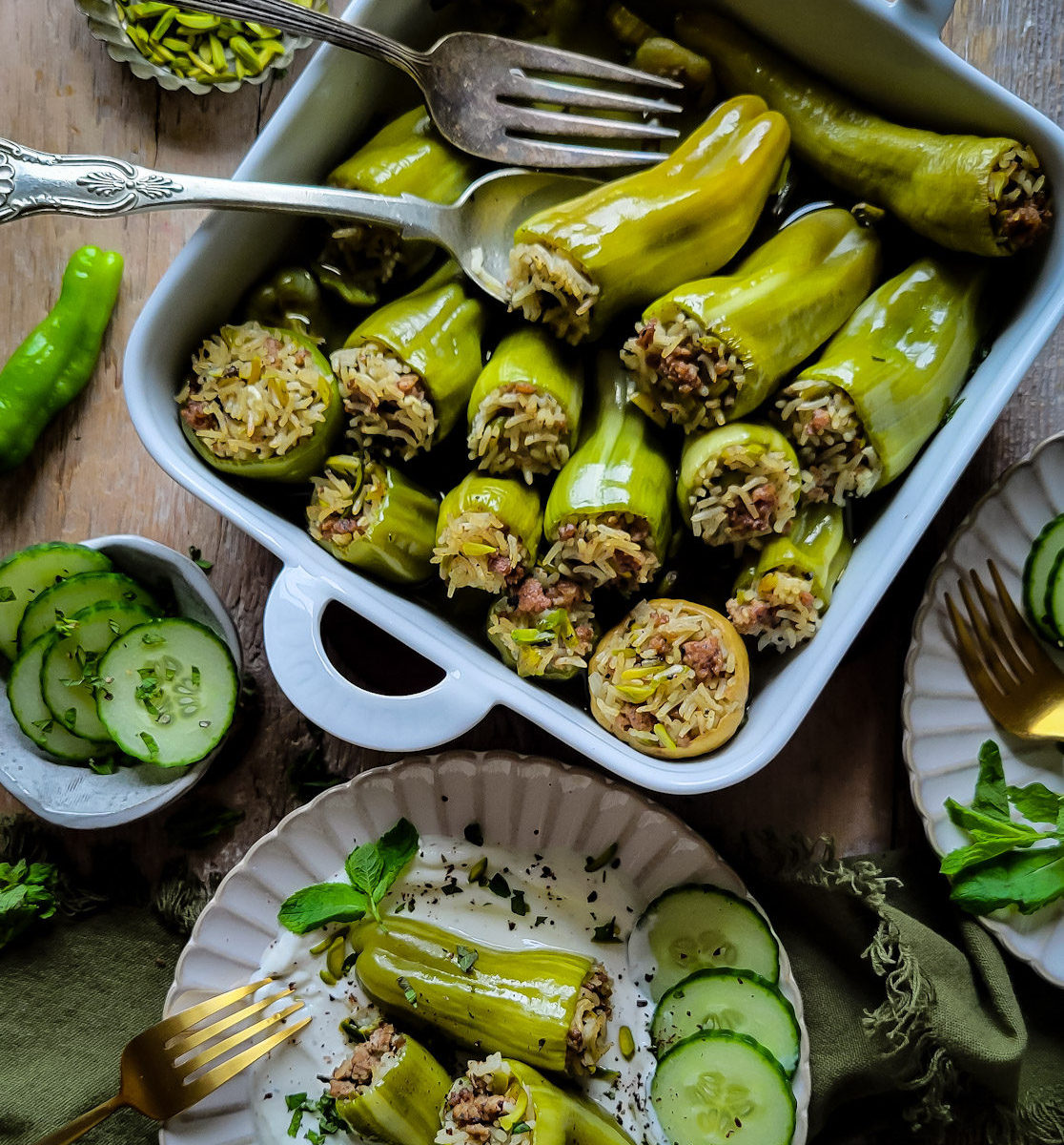 A baking dish filled with baked lamb and rice stuffed mini peppers. Nearby is a plate with cumin yogurt, three stuffed peppers, and cucumber slices. More cucumber slices, mint leaves and pistachios are on the side