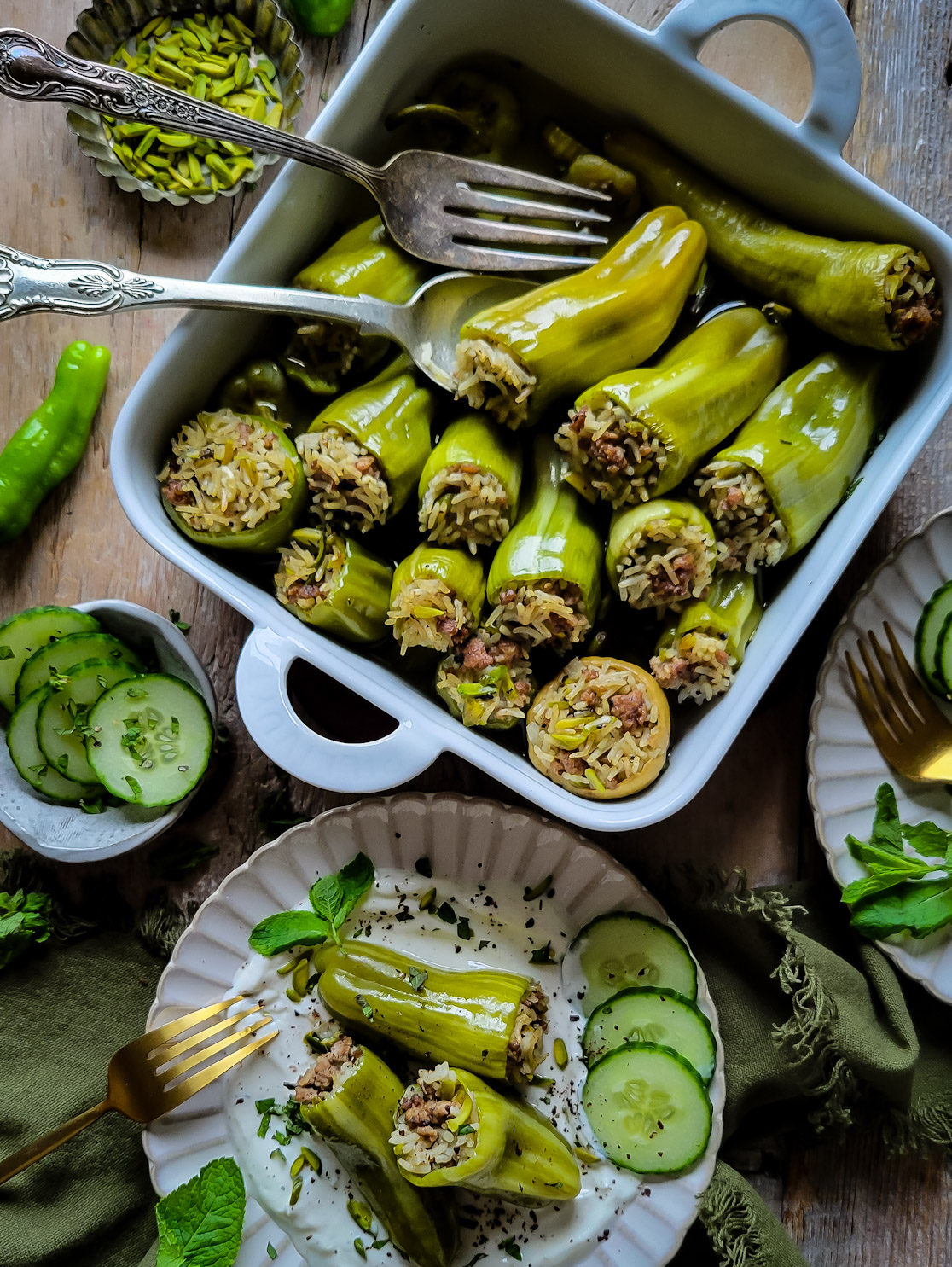 A baking dish filled with baked lamb and rice stuffed mini peppers. Nearby is a plate with cumin yogurt, three stuffed peppers, and cucumber slices. More cucumber slices, mint leaves and pistachios are on the side