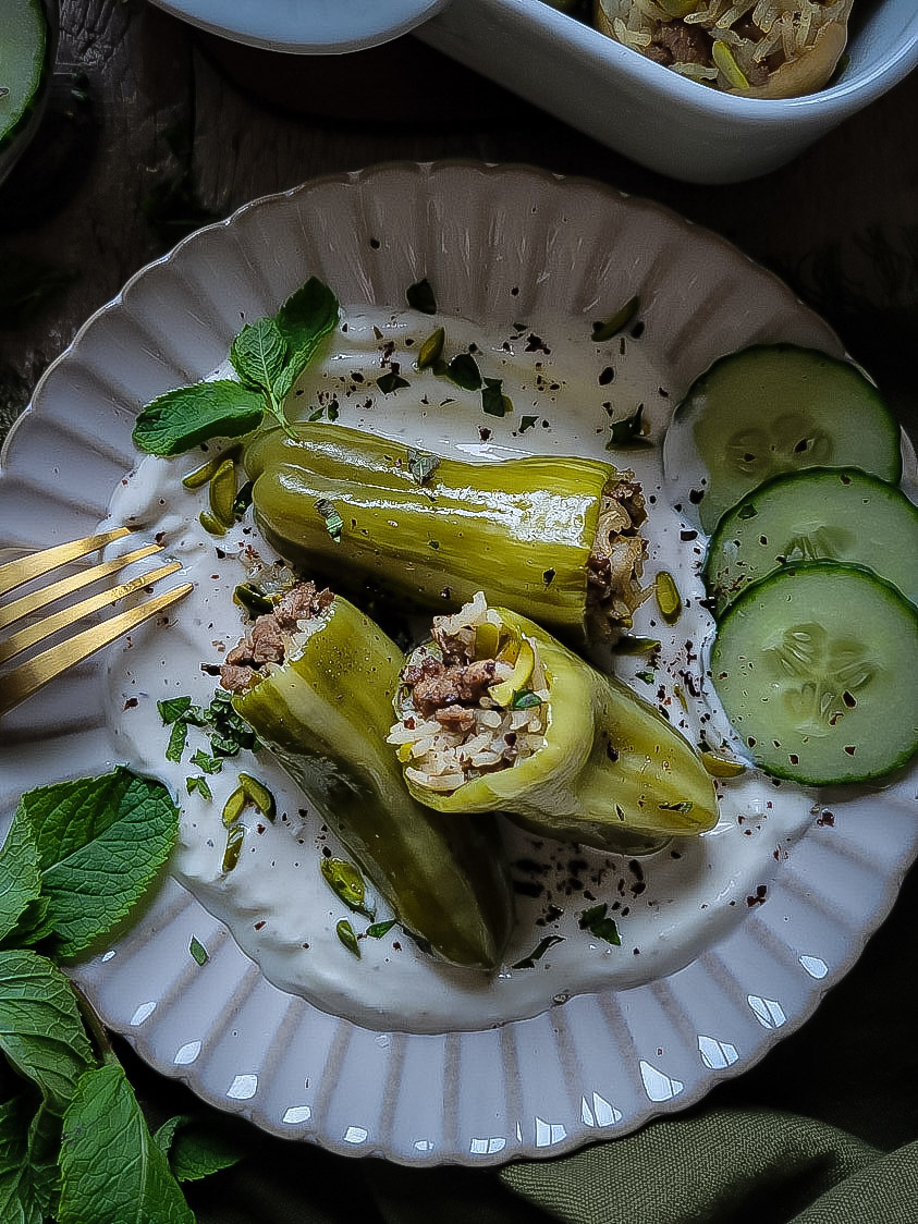 Three baked lamb and rice stuffed mini peppers sitting on a plate of cumin yogurt, cucumber slices, mint leaves and pistachios.